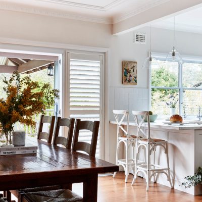 Premium Plantation Shutters 3 Interior photography of a fresh white Hamptons style kitchen with breakfast bar, cross back bar stools, polished floor boards and a rustic timber dining room setting to the left